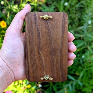 May include: A rectangular wooden plaque with a rich brown finish, featuring brass hardware at the top and bottom. The wood grain is visible, and the plaque is held in a hand against a blurred green background.