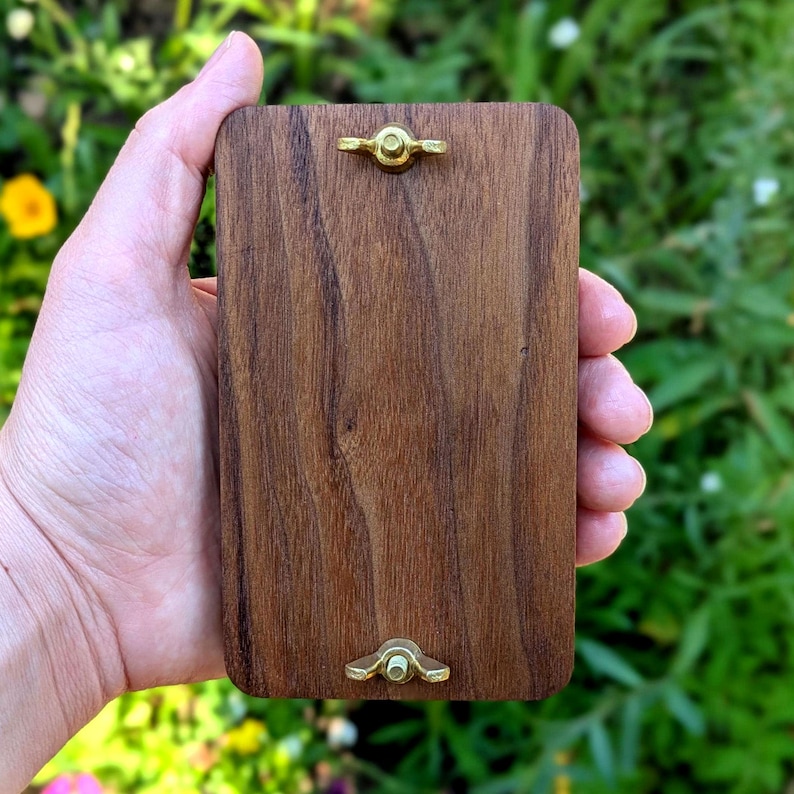 May include: A rectangular wooden plaque with a rich brown finish, featuring brass hardware at the top and bottom. The wood grain is visible, and the plaque is held in a hand against a blurred green background.