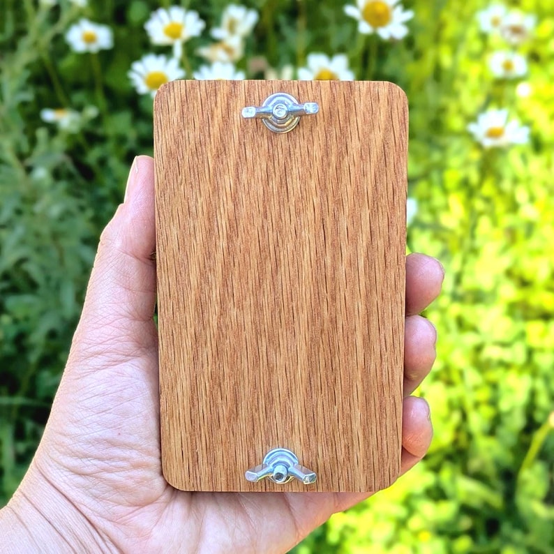 May include: A rectangular wooden plaque with a natural wood grain finish, secured with two silver-colored wing nuts. The plaque is held in a hand, with a blurred background of green foliage and white flowers.
