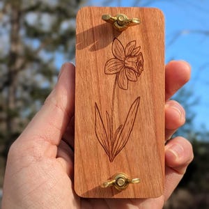 May include: A rectangular wooden craft with a daffodil flower etched into the surface. The wood is a warm reddish-brown color. Two brass wingnuts are attached to the top and bottom of the wood. The background is blurred.