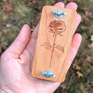 May include: A rectangular wooden plaque with a rose engraving. The plaque is light brown and has two silver-colored wing nuts attached. The rose is detailed with petals, leaves, and a stem. The background is blurred.