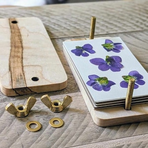May include: A wooden press for pressing flowers, with two brass wing nuts, two brass washers, and a stack of white cards with pressed purple flowers.