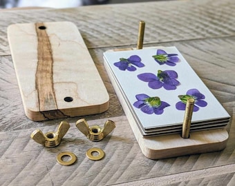 May include: A wooden press for pressing flowers, with two brass wing nuts, two brass washers, and a stack of white cards with pressed purple flowers.