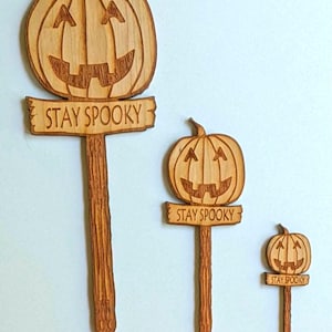 May include: Three wooden pumpkin signs with the text "Stay Spooky" on each. The signs are different sizes and are all carved with a pumpkin face.