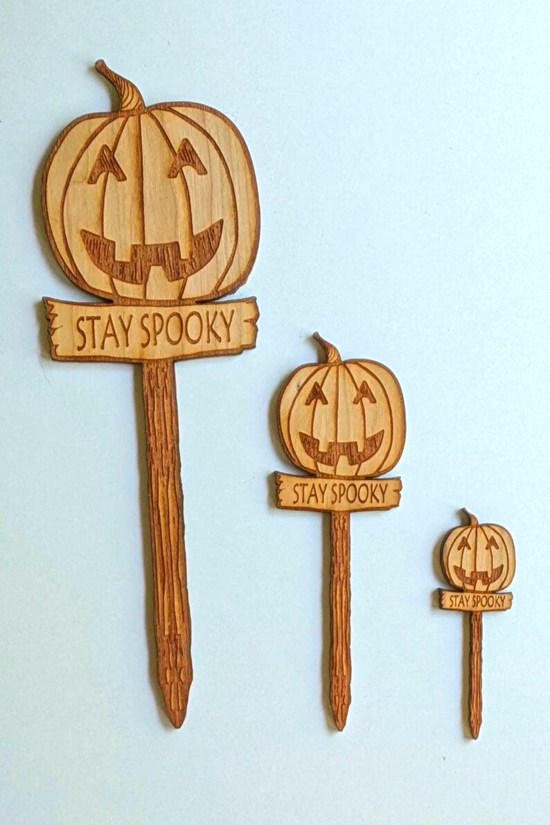 May include: Three wooden pumpkin signs with the text "Stay Spooky" on each. The signs are different sizes and are all carved with a pumpkin face.