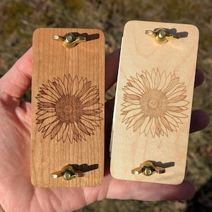 May include: Two wooden, rectangular items with brass hardware. One is light-colored, the other is darker. Each features a detailed sunflower engraving. The items are likely decorative or functional, possibly for a craft or home decor.