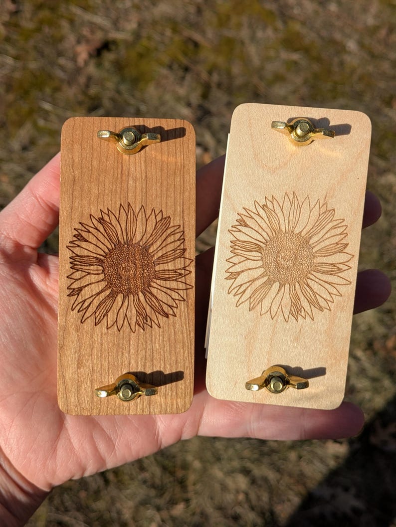 May include: Two wooden, rectangular items with brass hardware. One is light-colored, the other is darker. Each features a detailed sunflower engraving. The items are likely decorative or functional, possibly for a craft or home decor.