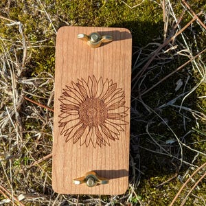 May include: A rectangular wooden board with a laser-etched sunflower design. The board has two brass wing nuts. The wood has a light brown color. The background is a mix of green moss and dry grass.