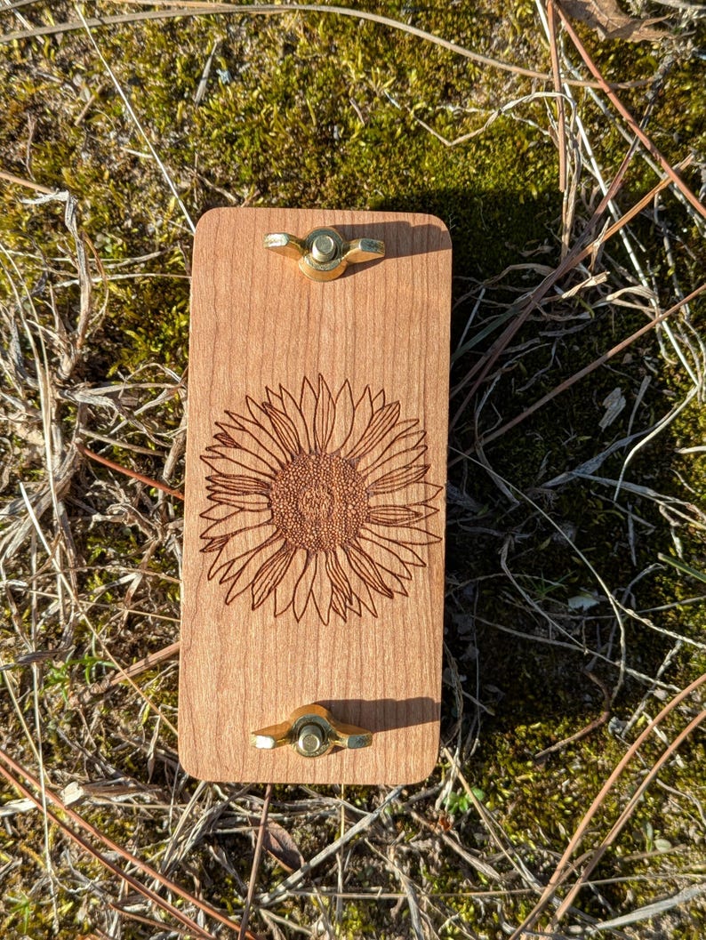 May include: A rectangular wooden board with a laser-etched sunflower design. The board has two brass wing nuts. The wood has a light brown color. The background is a mix of green moss and dry grass.