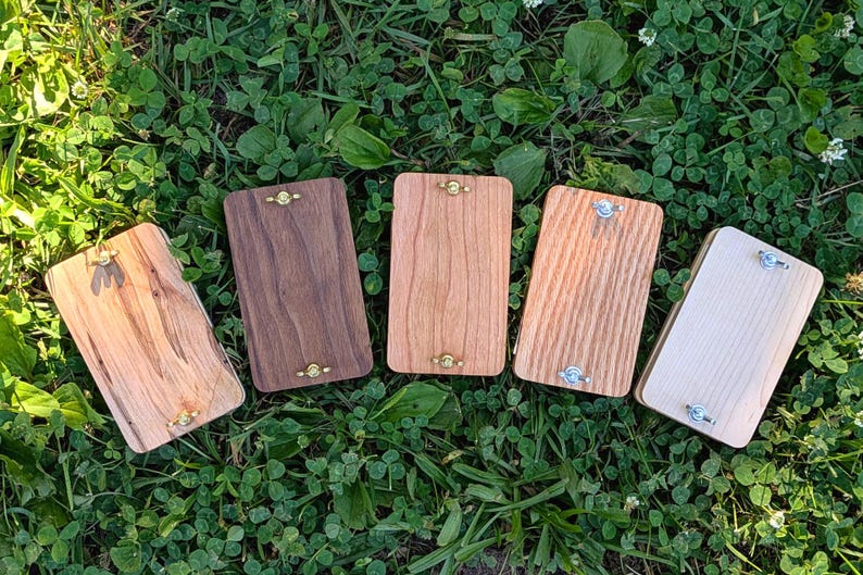 May include: Five rectangular wooden boards with varying wood grains and colors, arranged on a bed of green grass. Each board has a metal hinge and decorative hardware. The wood tones range from light to dark.