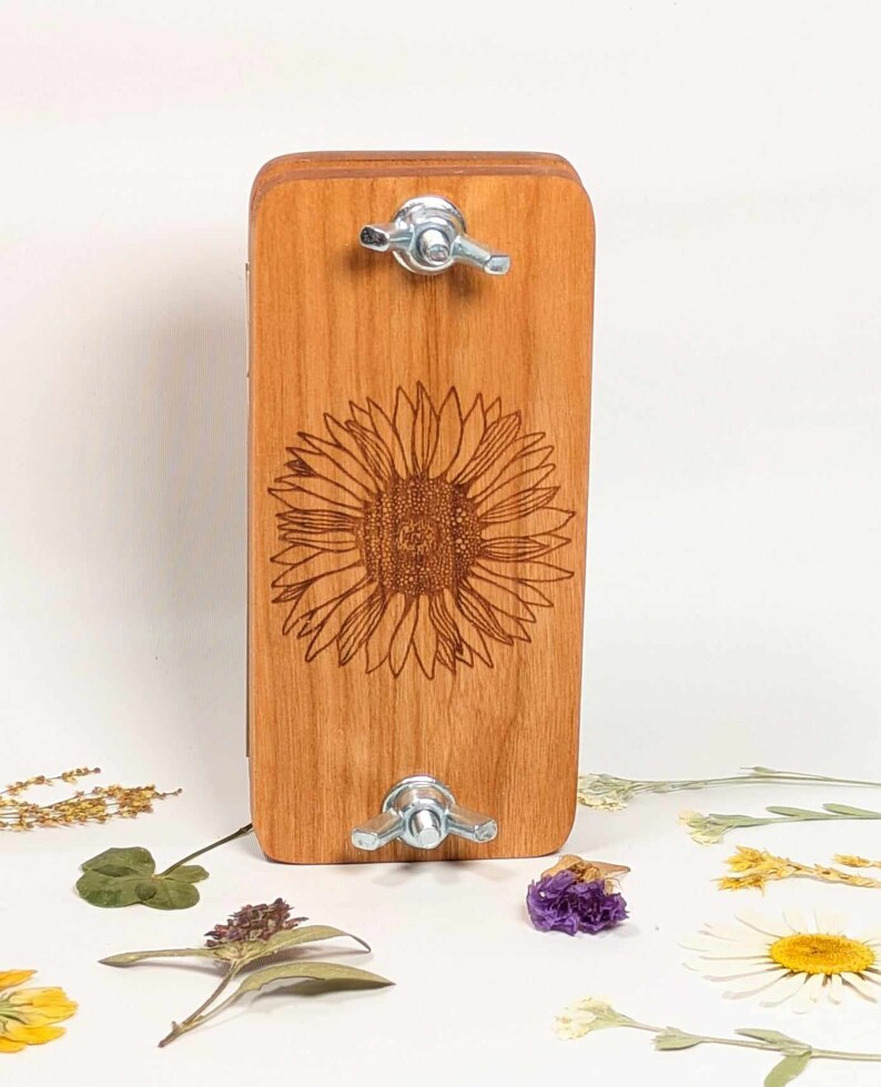May include: A wooden flower press with a sunflower design etched on the front. The press is rectangular with rounded edges and features two metal wingnuts for tightening. Dried flowers and foliage are scattered around the base.