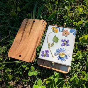 May include: A wooden flower press with a light brown finish, open to reveal pressed flowers in various colors. The flowers include yellow, pink, purple, and blue blooms, and green leaves. The press is lying on a bed of green grass.