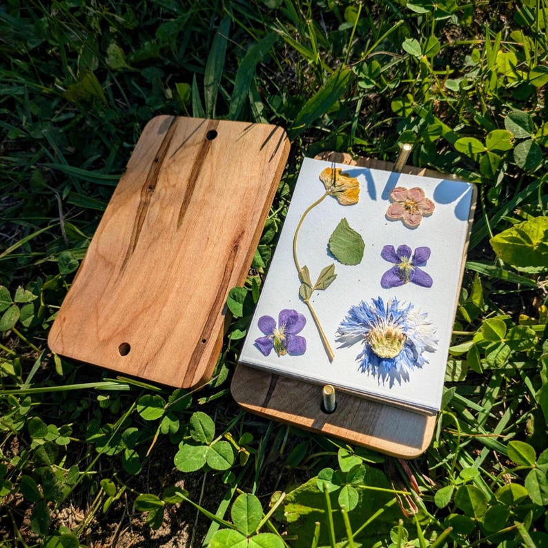 May include: A wooden flower press with a light brown finish, open to reveal pressed flowers in various colors. The flowers include yellow, pink, purple, and blue blooms, and green leaves. The press is lying on a bed of green grass.
