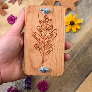 May include: A rectangular wooden leaf press with a detailed oak leaf design etched into the wood. The press has two metal wingnuts for securing items. The wood is light brown, and the design is a darker brown.