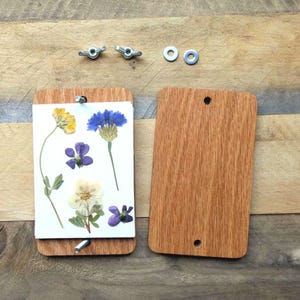 May include: A wooden flower press with pressed flowers. The press is made of two rectangular wooden boards, held together with wing nuts. Pressed flowers include yellow, blue, purple, and white blooms. The press is open, revealing the flowers.