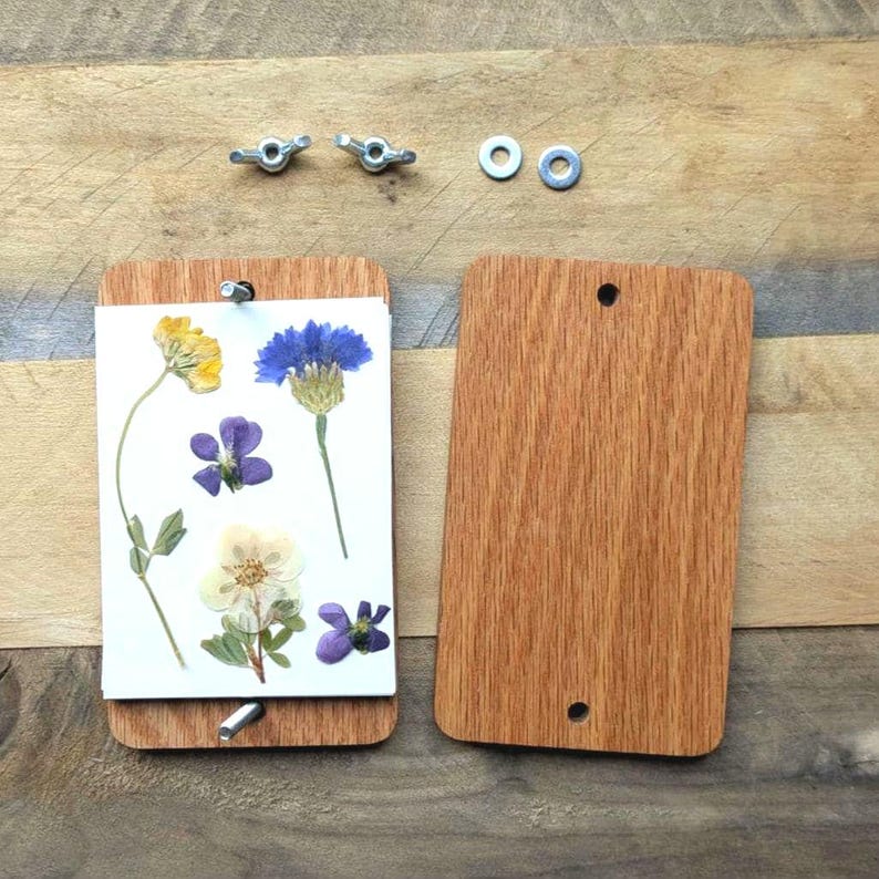 May include: A wooden flower press with pressed flowers. The press is made of two rectangular wooden boards, held together with wing nuts. Pressed flowers include yellow, blue, purple, and white blooms. The press is open, revealing the flowers.