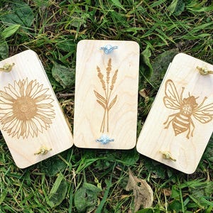 May include: Three wooden plant presses with engraved designs: a sunflower, wheat, and a bee. Each press has brass and silver hardware. The presses are rectangular and made of light-colored wood, resting on a bed of green grass.