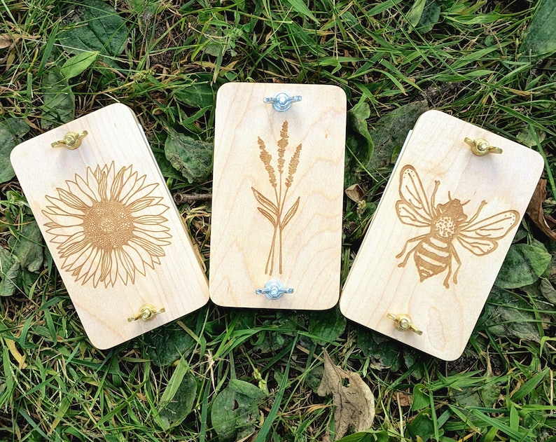 May include: Three wooden plant presses with engraved designs: a sunflower, wheat, and a bee. Each press has brass and silver hardware. The presses are rectangular and made of light-colored wood, resting on a bed of green grass.