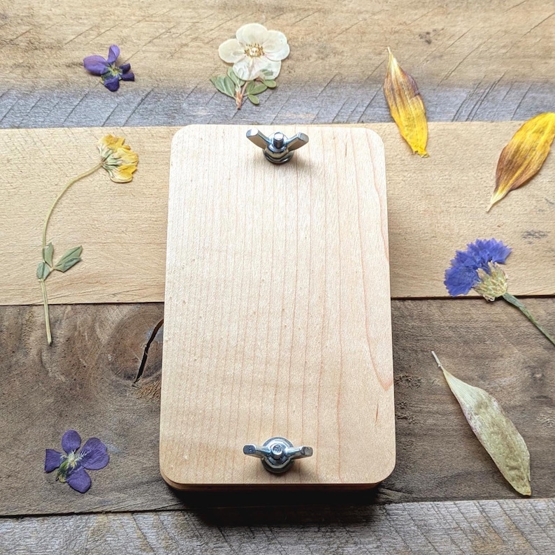 May include: A wooden flower press with two wing nuts, surrounded by dried flowers and leaves. The press is rectangular with rounded corners and a light wood finish. Various dried flowers and leaves are scattered around the press on a wooden surface.