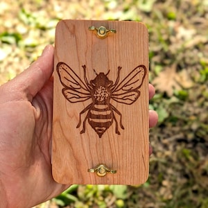 May include: A rectangular wooden box with a detailed engraved bee design. The box is light brown with brass hardware. The bee is depicted with intricate wing and body details.
