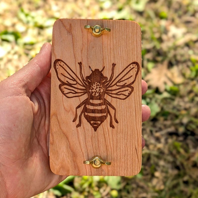 May include: A rectangular wooden box with a detailed engraved bee design. The box is light brown with brass hardware. The bee is depicted with intricate wing and body details.