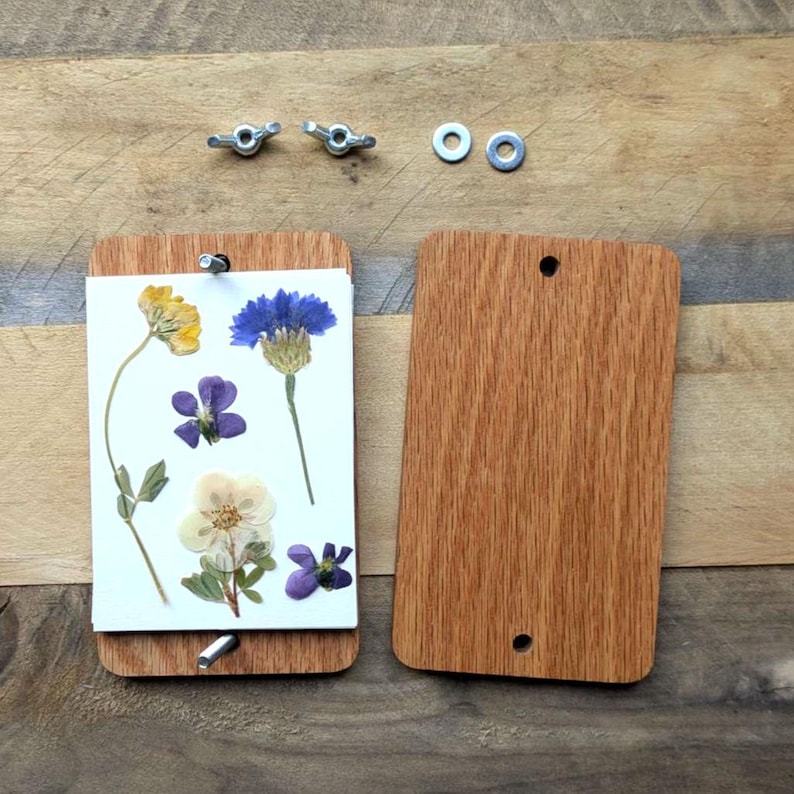 May include: A wooden flower press with pressed flowers. The press is made of two rectangular wooden boards, held together with wing nuts and bolts. Pressed flowers include yellow, blue, purple, and white blooms. The press is designed for preserving flowers.