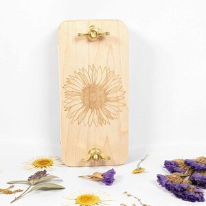 May include: A light-colored wooden flower press with a laser-etched sunflower design. The press has brass hardware and is surrounded by dried flowers, including daisies and purple statice.