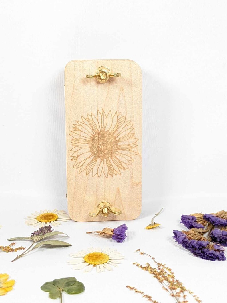 May include: A light-colored wooden flower press with a laser-etched sunflower design. The press has brass hardware and is surrounded by dried flowers, including daisies and purple statice.