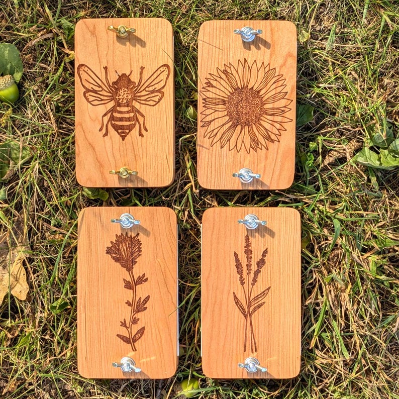 May include: Four wooden plant presses with engraved botanical designs: a bee, a sunflower, a flower, and a sprig of lavender. Each press has metal hardware for securing plants. The presses are rectangular and made of light-colored wood.