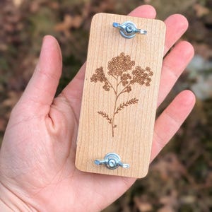 May include: A rectangular wooden plant press with a detailed floral design etched into the wood. The press is held together with two metal wing nuts. The wood has a light, natural color.