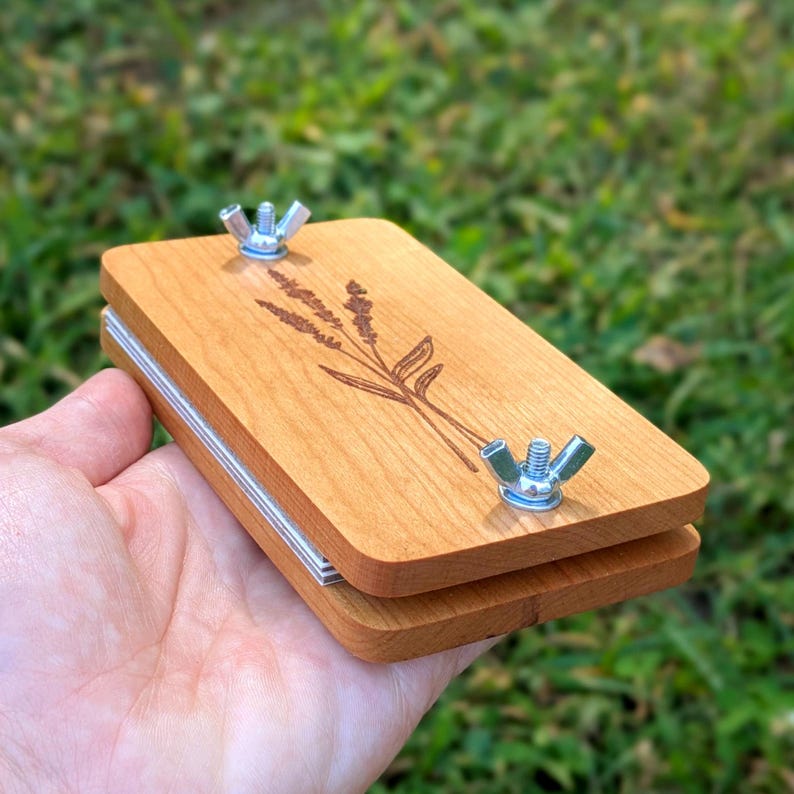 May include: A wooden flower press with a lavender design etched on the top. The press is made of light-colored wood and has two metal wing nuts for tightening. The press is held in a hand against a blurred green background.