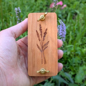 May include: A rectangular wooden plant press with a laser-etched lavender design. The press has brass hardware, including two wingnuts for tightening. The wood is a light brown color, and the design is a darker brown. The background is blurred green foliage.