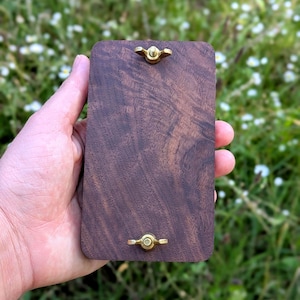 May include: A rectangular, dark brown wooden object with a brass butterfly nut at the top and bottom. The wood grain is visible. The object is held in a hand against a blurred green and white background.