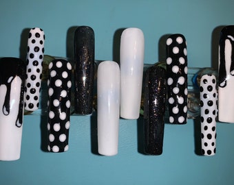 May include: A collection of ten artificial nails with diverse designs. The nails showcase black and white polka dots, solid black glitter, solid white, and a black dripping design on a white base. The nails are presented against a light blue backdrop.