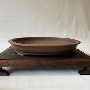 May include: A brown oval ceramic bonsai pot with a wooden stand. The stand has four legs and is made of dark wood.