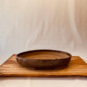 May include: A brown ceramic bonsai pot with four feet sits on a wooden tray. The pot has a slightly rounded shape and a textured surface.