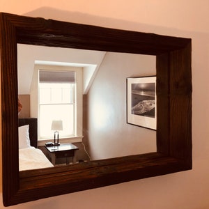 May include: A rectangular mirror with a dark brown, rustic wooden frame. The mirror reflects a room with a bed, a window with blinds, a lamp, and a framed black and white photograph. The frame has visible wood grain.