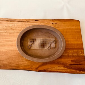 May include: A brown oval ceramic dish with a woven mesh insert sits on a wooden serving tray. The tray has a natural wood grain and a rough, uneven edge.