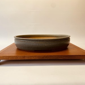 Peut inclure: Un bol en céramique grise à surface texturée repose sur un plateau rectangulaire en bois. Le plateau est en bois brun rougeâtre et présente un design simple et moderne. Le bol est de forme ovale et a un bord foncé.