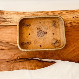 May include: A rectangular wooden tray with a natural wood grain finish. The tray has a smaller rectangular ceramic planter with a light brown glaze and a textured surface. The planter has four small holes and four black wire loops attached to the inside.