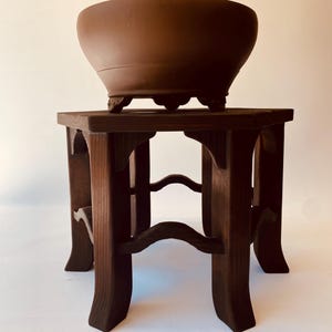May include: A matte brown ceramic bowl with a smooth, rounded rim and small decorative feet, resting on a dark brown wooden stand featuring six legs and curved support braces connecting the legs in a hexagonal arrangement.