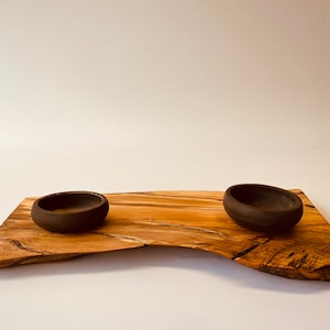 May include: Two small, dark brown ceramic bowls sit on a wooden serving tray with a natural, live edge finish. The tray has a light brown color with dark brown wood grain.