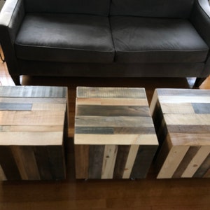 May include: Three wooden side tables with a rustic, reclaimed wood design. Each table is made of different shades of wood, including light brown, dark brown, and gray. The tables are arranged in a row, with the couch in the background.