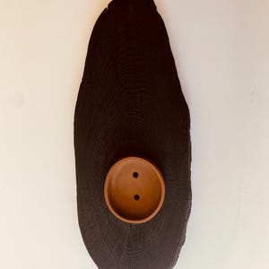 May include: A dark, oval-shaped wooden plaque with a circular, terracotta-colored bowl affixed to its center. The bowl has two small holes. The wood grain is visible, and the plaque is mounted on a white wall.