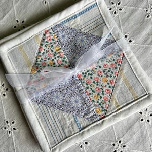 May include: A square quilted coaster set tied with a white ribbon. The coaster features a patchwork design with floral and striped fabric in shades of pink, blue, yellow, and white. The edges are finished with white binding.