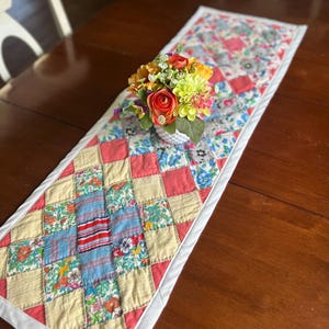 May include: A rectangular quilted table runner featuring a patchwork design of various floral and striped fabric squares in shades of pink, yellow, blue, and white, finished with a solid white border and topped with a small bouquet of artificial flowers.