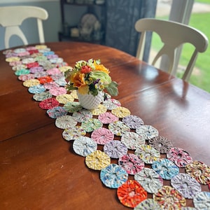 May include: A long table runner constructed from numerous small, circular fabric yo-yos in a variety of floral and solid patterns, featuring colors including yellow, pink, blue, red, and green, with a small white vase of flowers placed on top.