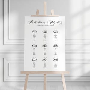 May include: A white seating chart on a wooden easel. The chart features elegant script and lists names under the years 2017 through 2025. The text "Finde deinen Sitzplatz" is at the top. The easel is made of light-colored wood.