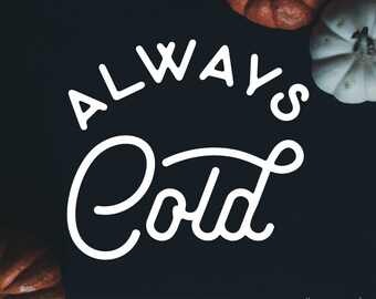 May include: White text on a black background reads "Always Cold" in a handwritten font. The text is surrounded by white and orange pumpkins.