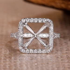 May include: A square-shaped engagement ring setting with a halo of small, sparkling stones. The ring features a crisscross design in the centre, designed to hold a gemstone. The band is also adorned with small stones, adding to its brilliance.